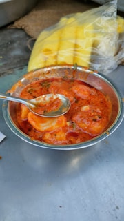 A bowl filled with a spicy, red curry containing pieces of food, possibly boiled eggs, and garnished with herbs. A spoon rests inside the bowl. In the background, there is a packaged, sliced yellow food item, possibly pineapples, placed on a woven surface.