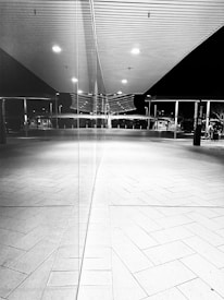 A black and white image featuring a modern architectural setting with clean lines and reflective surfaces. The scene is split symmetrically by a large glass window in the center, creating a mirror effect. The ceiling is composed of horizontal panels, with evenly spaced lights that cast a stark contrast with shadows. The floor is tiled with a geometric pattern, contributing to the overall minimalist aesthetic. In the background, structural elements like pillars and an open area can be seen, possibly indicating a public or commercial space.