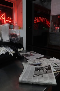 A dimly lit counter with several newspapers spread out, including the Los Angeles Times. A plastic container holds various condiment packets, and there is a neon red light reflecting in the window. The surface is metallic, and there appears to be a clutter of items such as paper towels and napkins.