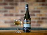 A bottle of Pinot Grigio and a glass filled with white wine are placed on a wooden surface. The background consists of a blurred brick wall, adding a rustic feel to the scene. The label on the bottle indicates it is from South Eastern Australia, vintage 2022.