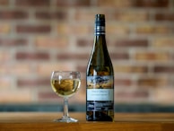A bottle of Pinot Grigio and a glass filled with white wine are placed on a wooden surface. The background consists of a blurred brick wall, adding a rustic feel to the scene. The label on the bottle indicates it is from South Eastern Australia, vintage 2022.