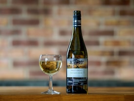 A bottle of Pinot Grigio and a glass filled with white wine are placed on a wooden surface. The background consists of a blurred brick wall, adding a rustic feel to the scene. The label on the bottle indicates it is from South Eastern Australia, vintage 2022.