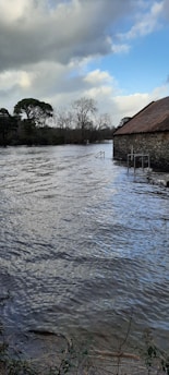 A stone building with a sloped roof appears partially submerged in floodwaters that extend across the scene. The water is rippling, reflecting the grey and blue coloration of the sky. Trees with bare branches and evergreens are visible in the background under a cloudy sky, with some patches of blue and sunlight breaking through.
