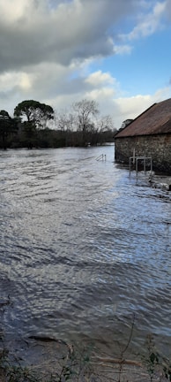 A stone building with a sloped roof appears partially submerged in floodwaters that extend across the scene. The water is rippling, reflecting the grey and blue coloration of the sky. Trees with bare branches and evergreens are visible in the background under a cloudy sky, with some patches of blue and sunlight breaking through.