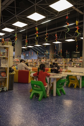 A library or reading area with colorful seating arrangements is filled with children and adults. The ceiling features hanging decorative spirals in various colors. Bookshelves and tables are scattered around, with people engaging in reading or activities.