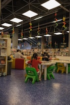 A library or reading area with colorful seating arrangements is filled with children and adults. The ceiling features hanging decorative spirals in various colors. Bookshelves and tables are scattered around, with people engaging in reading or activities.
