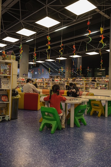 A library or reading area with colorful seating arrangements is filled with children and adults. The ceiling features hanging decorative spirals in various colors. Bookshelves and tables are scattered around, with people engaging in reading or activities.
