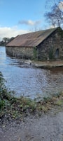 A rustic stone building with a rusty metal roof is partially submerged in floodwaters. The surrounding area shows trees and bushes with patches of greenery. The sky is clear with some clouds, casting reflections on the calm water surface.