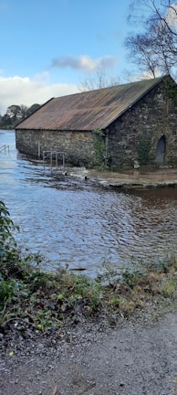 A rustic stone building with a rusty metal roof is partially submerged in floodwaters. The surrounding area shows trees and bushes with patches of greenery. The sky is clear with some clouds, casting reflections on the calm water surface.