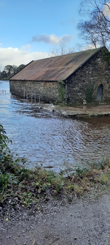 A rustic stone building with a rusty metal roof is partially submerged in floodwaters. The surrounding area shows trees and bushes with patches of greenery. The sky is clear with some clouds, casting reflections on the calm water surface.