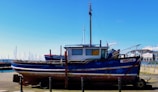 A blue and white boat with some rust marks is docked at a marina. The boat has a 'For Sale' sign displayed in the window. In the background, there are more boats with masts and a clear blue sky. Buildings can be seen on the right side of the image.