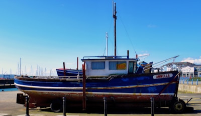 A blue and white boat with some rust marks is docked at a marina. The boat has a 'For Sale' sign displayed in the window. In the background, there are more boats with masts and a clear blue sky. Buildings can be seen on the right side of the image.
