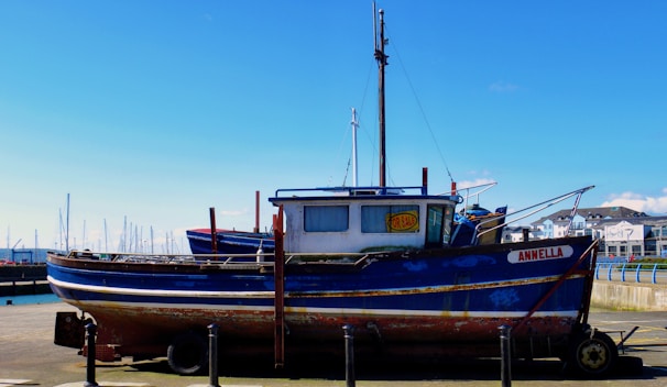 A blue and white boat with some rust marks is docked at a marina. The boat has a 'For Sale' sign displayed in the window. In the background, there are more boats with masts and a clear blue sky. Buildings can be seen on the right side of the image.