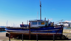 A blue and white boat with some rust marks is docked at a marina. The boat has a 'For Sale' sign displayed in the window. In the background, there are more boats with masts and a clear blue sky. Buildings can be seen on the right side of the image.