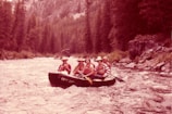 Four individuals are enjoying a rafting adventure on a swift river surrounded by dense forest. They are seated in a rubber raft, wearing life jackets and hats, indicating a focus on safety and outdoor fun. The river is turbulent, creating a sense of motion and excitement.