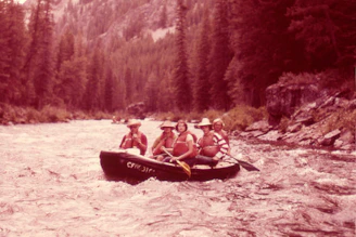 Four individuals are enjoying a rafting adventure on a swift river surrounded by dense forest. They are seated in a rubber raft, wearing life jackets and hats, indicating a focus on safety and outdoor fun. The river is turbulent, creating a sense of motion and excitement.