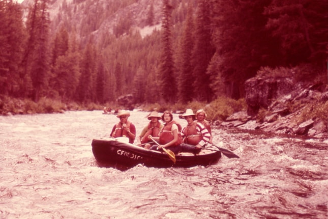 Four individuals are enjoying a rafting adventure on a swift river surrounded by dense forest. They are seated in a rubber raft, wearing life jackets and hats, indicating a focus on safety and outdoor fun. The river is turbulent, creating a sense of motion and excitement.