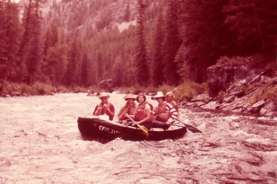 Four individuals are enjoying a rafting adventure on a swift river surrounded by dense forest. They are seated in a rubber raft, wearing life jackets and hats, indicating a focus on safety and outdoor fun. The river is turbulent, creating a sense of motion and excitement.