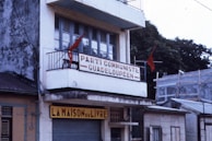A building with a weathered exterior features a balcony adorned with red flags. The sign on the balcony reads 'Parti Communiste Guadeloupeen,' reflecting a political theme. Below, a shop called 'La Maison du Livre' is visible, suggesting a bookstore or related business. The building is surrounded by other structures and partially obscured trees.