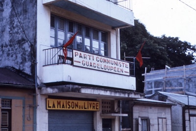 A building with a weathered exterior features a balcony adorned with red flags. The sign on the balcony reads 'Parti Communiste Guadeloupeen,' reflecting a political theme. Below, a shop called 'La Maison du Livre' is visible, suggesting a bookstore or related business. The building is surrounded by other structures and partially obscured trees.