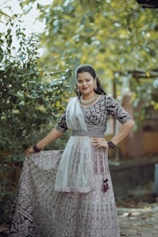 A woman is dressed in an intricately designed traditional outfit featuring a richly embroidered pattern. She stands outdoors amidst lush greenery, which provides a natural, vibrant backdrop. Her attire is complemented by elegant jewelry, including a necklace and earrings, and she wears matching glass bangles. The fabric of her dress has a detailed lace overlay and is held gracefully in her hand.