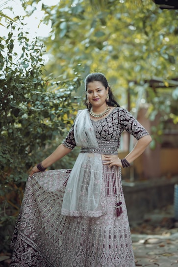 A woman is dressed in an intricately designed traditional outfit featuring a richly embroidered pattern. She stands outdoors amidst lush greenery, which provides a natural, vibrant backdrop. Her attire is complemented by elegant jewelry, including a necklace and earrings, and she wears matching glass bangles. The fabric of her dress has a detailed lace overlay and is held gracefully in her hand.