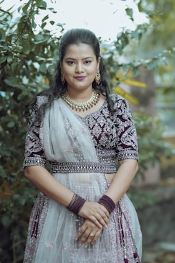 A woman is standing outdoors in front of foliage. She is wearing traditional attire, adorned with intricate embroidery and a sheer, embellished dupatta. She has her hands clasped in front of her. Her hair is styled neatly, and she is wearing jewelry including earrings and a necklace.