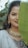 A woman with long, dark hair partially obscured by a large green leaf. She wears traditional jewelry including a necklace with pearls and beads, and ornate earrings. Her attire features intricate embroidery with a mix of dark and light colors. The background is blurred foliage.