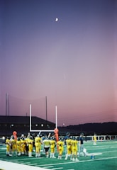 A group of young football players in yellow jerseys gather on a sports field under the evening sky. The goalposts and training equipment are visible, while the moon shines brightly in a clear dusk sky, creating a serene backdrop.