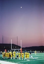 A group of young football players in yellow jerseys gather on a sports field under the evening sky. The goalposts and training equipment are visible, while the moon shines brightly in a clear dusk sky, creating a serene backdrop.