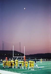 A group of young football players in yellow jerseys gather on a sports field under the evening sky. The goalposts and training equipment are visible, while the moon shines brightly in a clear dusk sky, creating a serene backdrop.