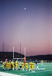 A group of young football players in yellow jerseys gather on a sports field under the evening sky. The goalposts and training equipment are visible, while the moon shines brightly in a clear dusk sky, creating a serene backdrop.
