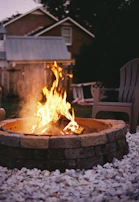 A warm fire burns brightly within a stone fire pit surrounded by a bed of white pebbles. A few outdoor chairs are placed nearby under the subdued light of early evening, with a backdrop of a wooden fence and a house.