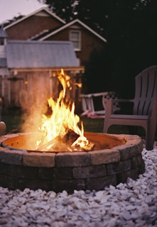A warm fire burns brightly within a stone fire pit surrounded by a bed of white pebbles. A few outdoor chairs are placed nearby under the subdued light of early evening, with a backdrop of a wooden fence and a house.