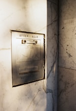An old-fashioned metallic after-hours deposit slot is embedded in a marble wall. The slot has text indicating it's for envelopes, and there are visible screws and a lock mechanism. Sunlight casts soft shadows across the surface, highlighting the aged texture of the metal and the veining in the marble.