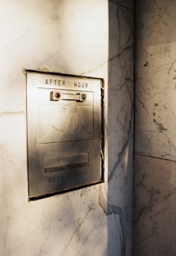 An old-fashioned metallic after-hours deposit slot is embedded in a marble wall. The slot has text indicating it's for envelopes, and there are visible screws and a lock mechanism. Sunlight casts soft shadows across the surface, highlighting the aged texture of the metal and the veining in the marble.
