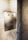 An old-fashioned metallic after-hours deposit slot is embedded in a marble wall. The slot has text indicating it's for envelopes, and there are visible screws and a lock mechanism. Sunlight casts soft shadows across the surface, highlighting the aged texture of the metal and the veining in the marble.