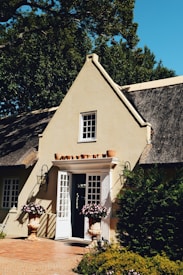 A charming cottage with a thatched roof and a light-colored facade. The entrance features white French doors adorned with potted plants on each side. Several terra cotta pots are arranged above the doorway. Greenery surrounds the house, and a clear blue sky is visible.