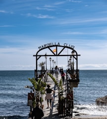 A wooden pier extends over a calm sea with several people walking along it. The entrance is marked by an archway with 'Marbella Club' written on it, and a Spanish flag is mounted on top. Palm trees and lampposts line the pier, creating a scenic walkway. The sky above is clear with subtle wispy clouds, suggesting a pleasant day.