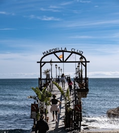 A wooden pier extends over a calm sea with several people walking along it. The entrance is marked by an archway with 'Marbella Club' written on it, and a Spanish flag is mounted on top. Palm trees and lampposts line the pier, creating a scenic walkway. The sky above is clear with subtle wispy clouds, suggesting a pleasant day.