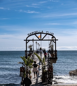 A wooden pier extends over a calm sea with several people walking along it. The entrance is marked by an archway with 'Marbella Club' written on it, and a Spanish flag is mounted on top. Palm trees and lampposts line the pier, creating a scenic walkway. The sky above is clear with subtle wispy clouds, suggesting a pleasant day.