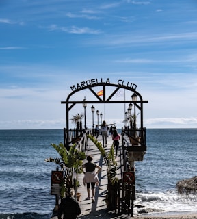 A wooden pier extends over a calm sea with several people walking along it. The entrance is marked by an archway with 'Marbella Club' written on it, and a Spanish flag is mounted on top. Palm trees and lampposts line the pier, creating a scenic walkway. The sky above is clear with subtle wispy clouds, suggesting a pleasant day.