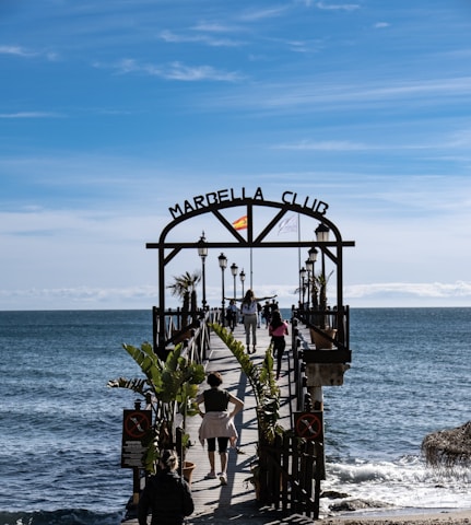 A wooden pier extends over a calm sea with several people walking along it. The entrance is marked by an archway with 'Marbella Club' written on it, and a Spanish flag is mounted on top. Palm trees and lampposts line the pier, creating a scenic walkway. The sky above is clear with subtle wispy clouds, suggesting a pleasant day.