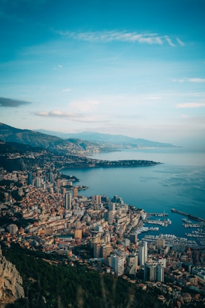 The image captures a panoramic view of a coastal city with dense urban development, featuring tall buildings and a bustling marina. The city extends along a rugged coastline with clear blue waters. Hills or mountains can be seen in the background under a partially cloudy sky.