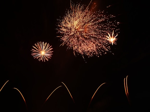 Several bright fireworks explode against a dark sky, with bursts of orange and red creating intricate patterns. Smaller fireworks can be seen in the foreground with trails leading up to the explosions.
