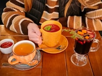 A person wearing a striped sweater holds an orange cup filled with a red beverage garnished with basil. On the table, there is a cup of tea in a speckled mug, a small dish of sauce, and a clear glass mug containing a dark drink with assorted fruit and herbs.