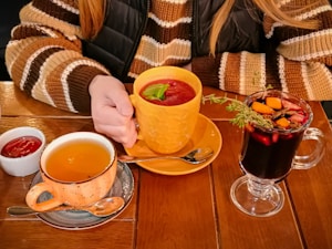 A person wearing a striped sweater holds an orange cup filled with a red beverage garnished with basil. On the table, there is a cup of tea in a speckled mug, a small dish of sauce, and a clear glass mug containing a dark drink with assorted fruit and herbs.