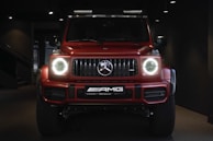 A red, powerful SUV with distinctive front headlights is displayed in a dimly lit showroom setting. The vehicle features a prominent grille with a recognizable emblem at the center and has a sleek, modern design that underscores its luxury and performance attributes.