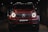 A red, powerful SUV with distinctive front headlights is displayed in a dimly lit showroom setting. The vehicle features a prominent grille with a recognizable emblem at the center and has a sleek, modern design that underscores its luxury and performance attributes.