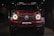 A red, powerful SUV with distinctive front headlights is displayed in a dimly lit showroom setting. The vehicle features a prominent grille with a recognizable emblem at the center and has a sleek, modern design that underscores its luxury and performance attributes.